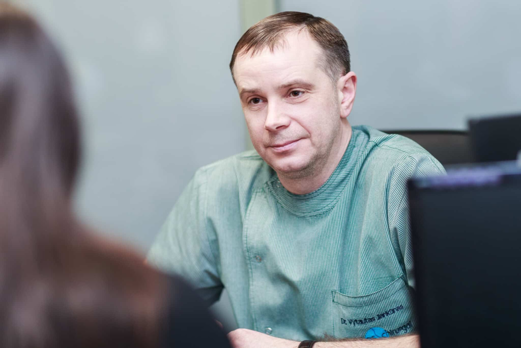 Vyras gydytojas žalia medicinine uniforma sėdi prie stalo ir atidžiai klausosi paciento. Nuotrauka primena medicinos kabinetą Lietuvoje, atspindintį 2025 m. plastinės chirurgijos tendencijas.